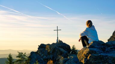 Eine Frau sitzt mit dem Rücken zur Kamera auf einem Feld. Sie blickt in Richtung des Gipfelkreuzes vom Silberberg. | © Bodenmais Tourismus & Marketing GmbH