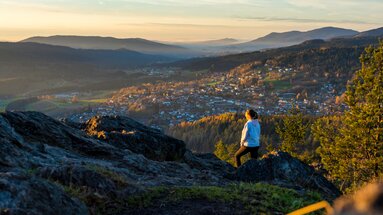 Ein Frau steht mit dem Rücken zur Kamera und blickt vom Silberberg aus hinab nach Bodenmais. Das herbstliche Tal wird von der untergehenden Sonne angestrahlt. | © Bodenmais Tourismus & Marketing GmbH